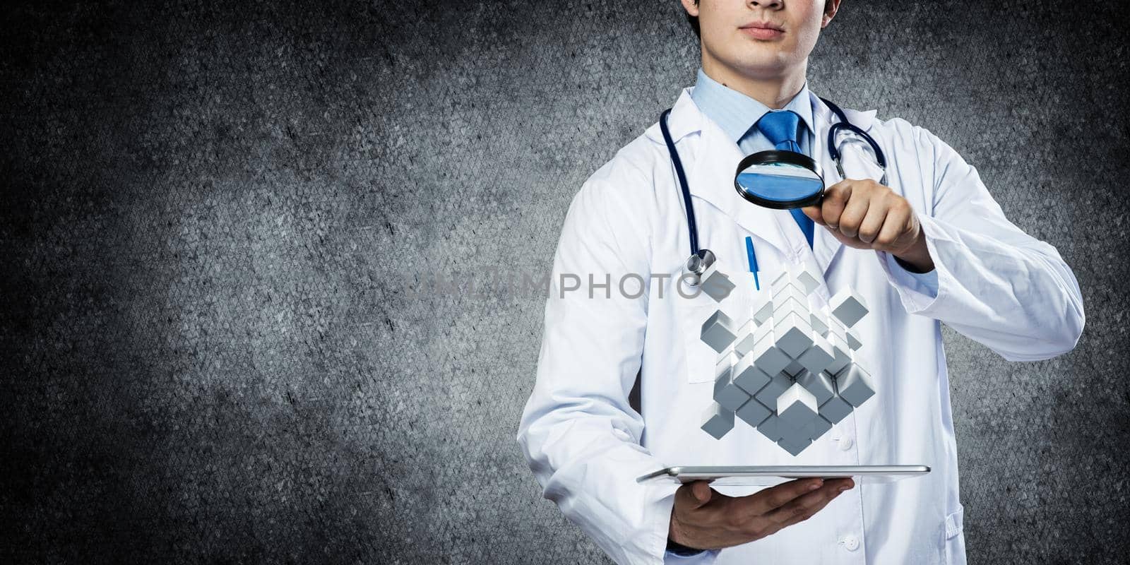 Horizontal shot of young doctor in white medical uniform studying multiple cubes in his hands while standing against gray dark wall on background.