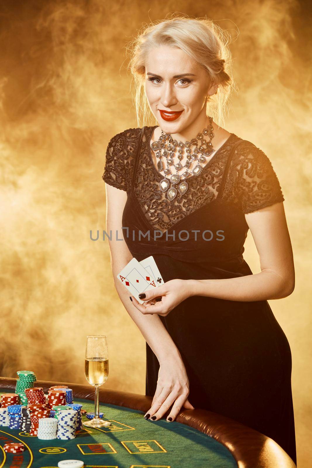 A woman in a black dress with cards in her hands is standing near the poker table. On the poker table are stacks of colored chips and a glass of champagne