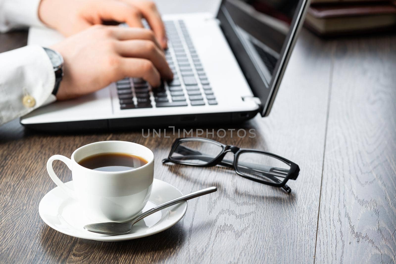 Businessman sitting at desk and working with laptop computer. Close-up of man hands typing on keyboard in office. Businessman workplace with glasses and cup of coffee. Innovations and entrepreneurship