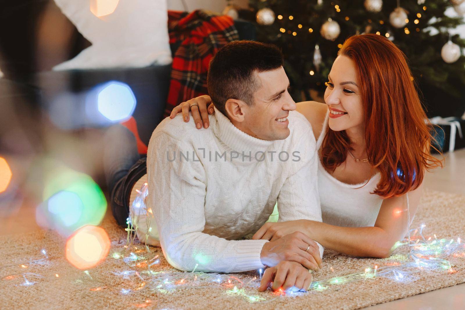 a happy married couple is lying on the floor near the Christmas tree at home.