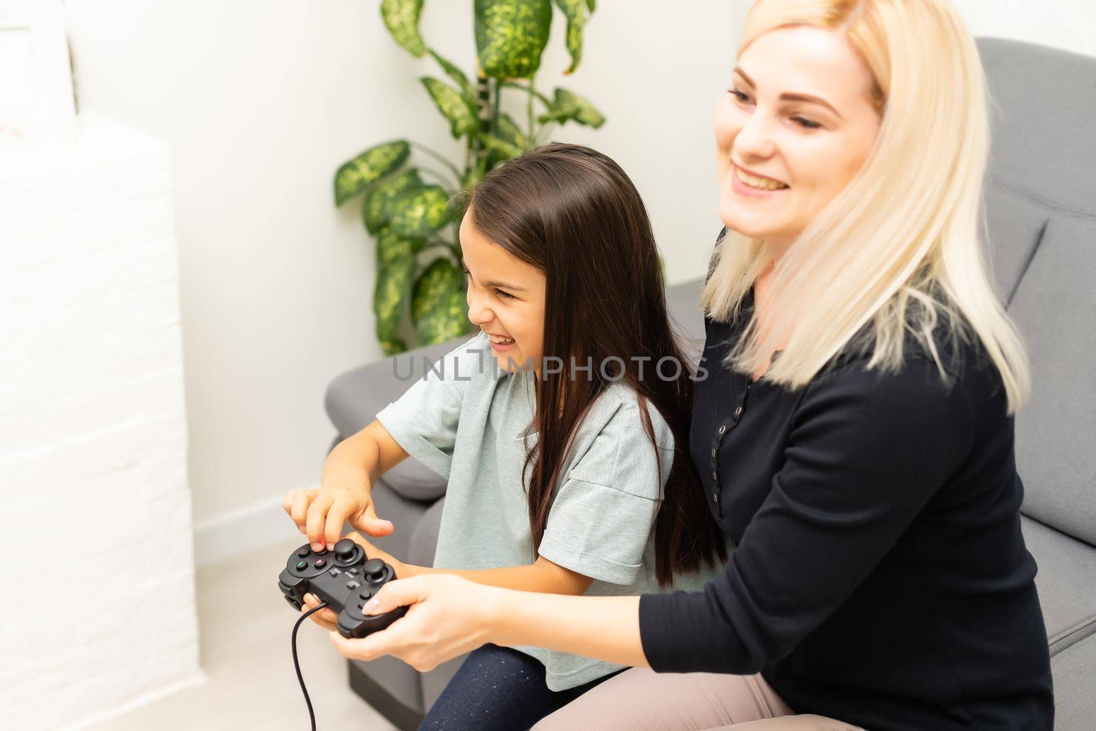 good relationship cute little girl with young mother using joystick playing video game sitting together in living room enjoying family holiday. by Andelov13