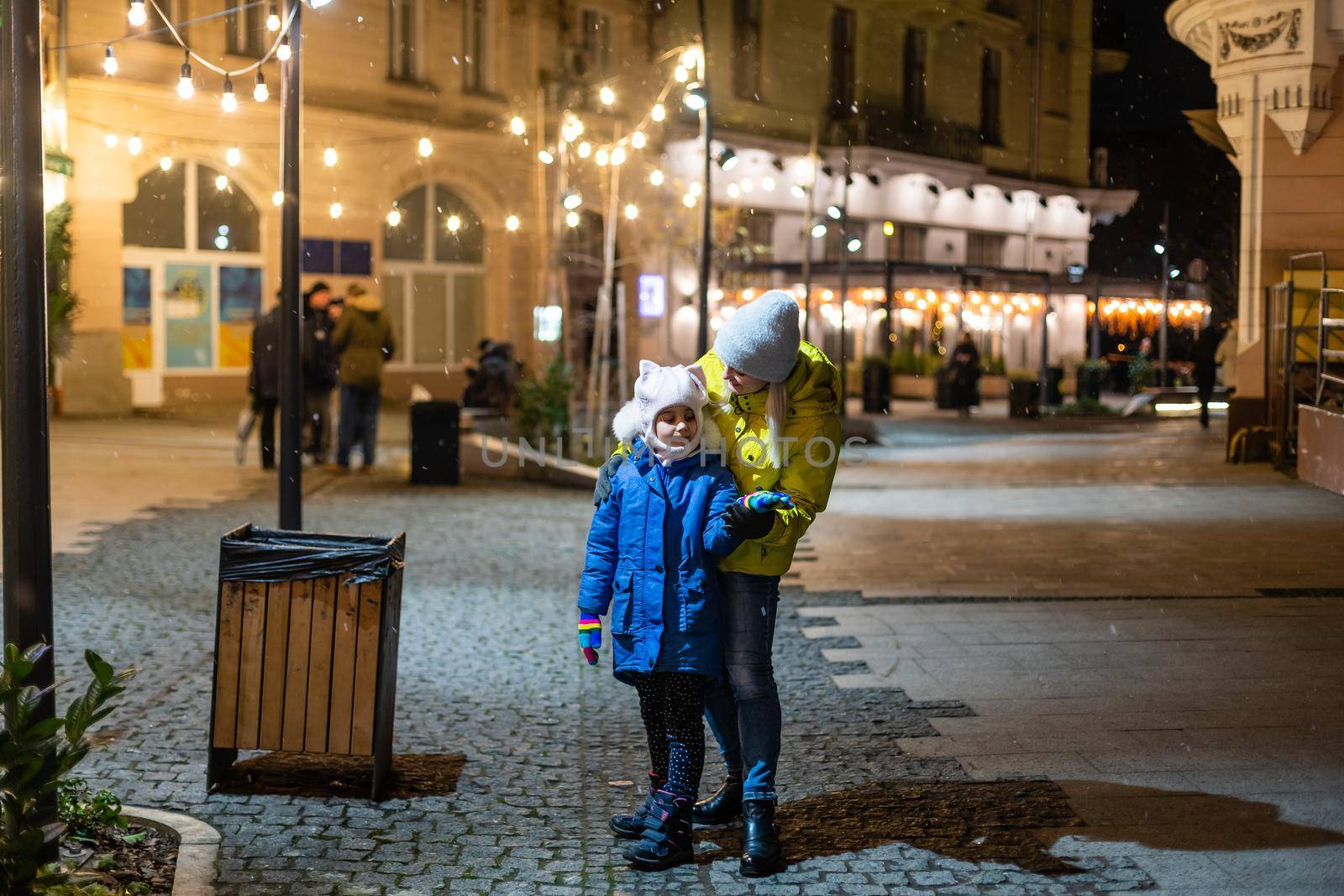 Mother embracing daughter while standing on the street in front of store window.