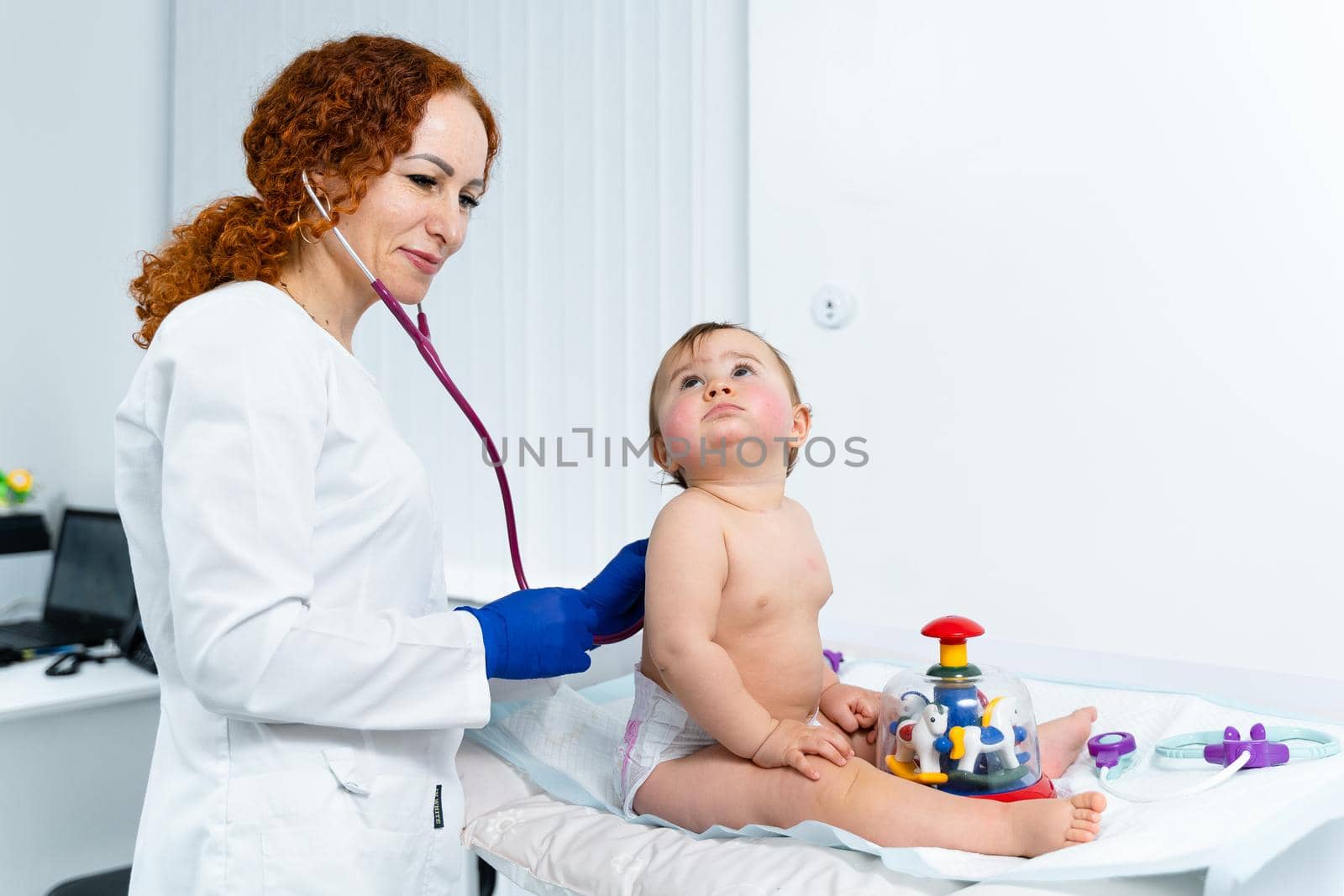 Red-haired female doctor examines little child for less than a year in a modern clinic. Pediatrician during examination baby in the hospital. Childrens healthcare. Diagnostics and treatment for kid by Tomashevska