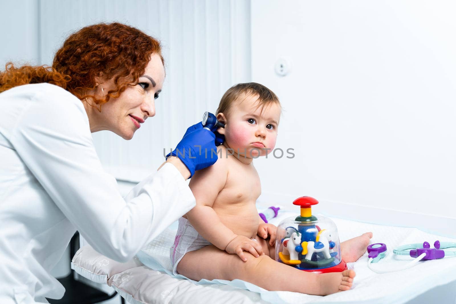 Red-haired female doctor examines little child for less than a year in a modern clinic. Pediatrician during examination baby in the hospital. Childrens healthcare. Diagnostics and treatment for kid.