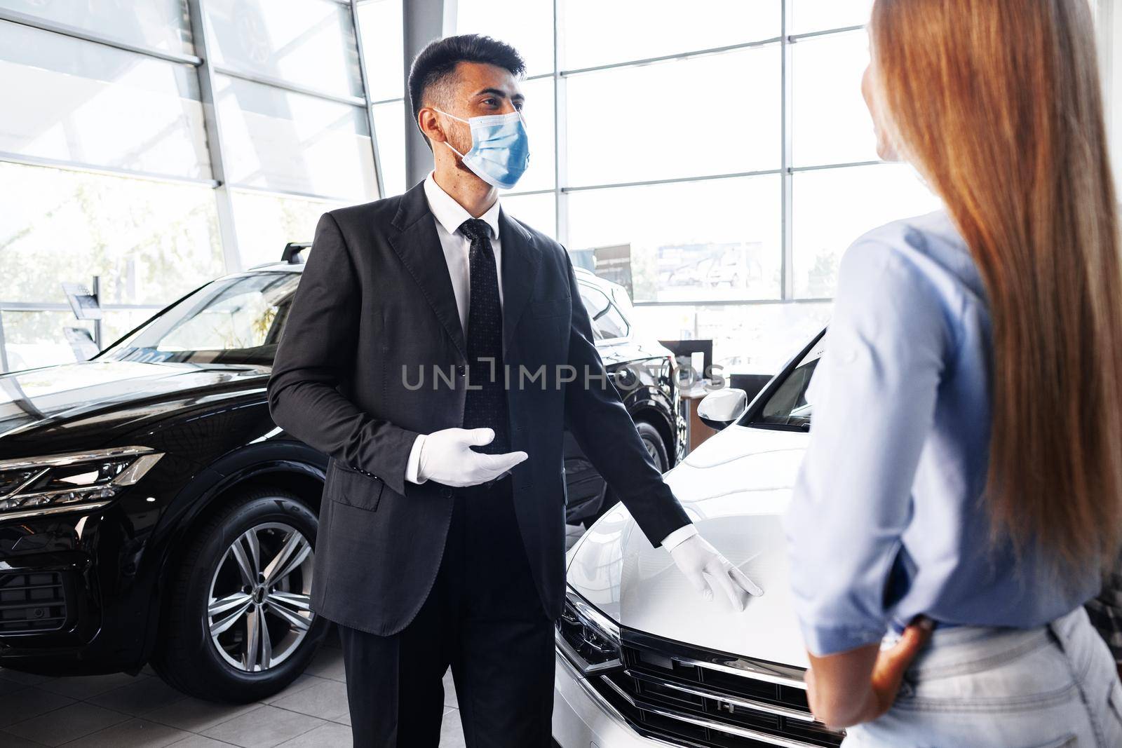 Man car salesman in face mask talking to a client in car showroom