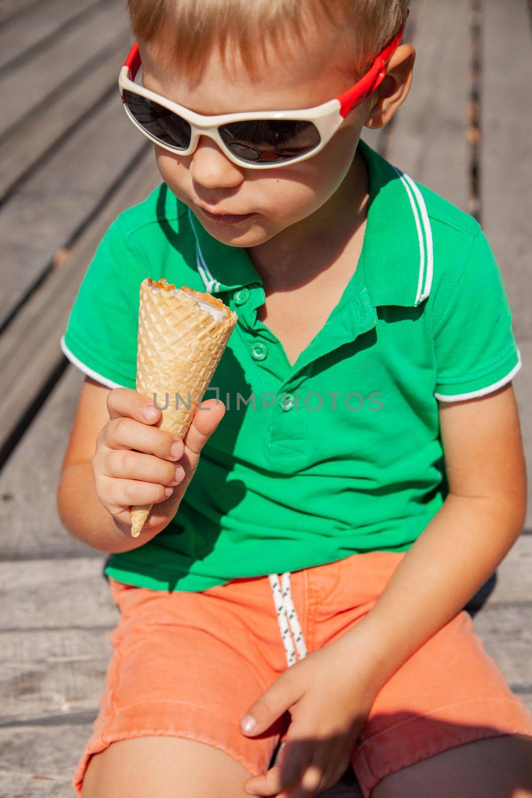 Cute little boy in stylish sunglasses eating yummy vanilla ice cream cone and enjoying summertime in park
