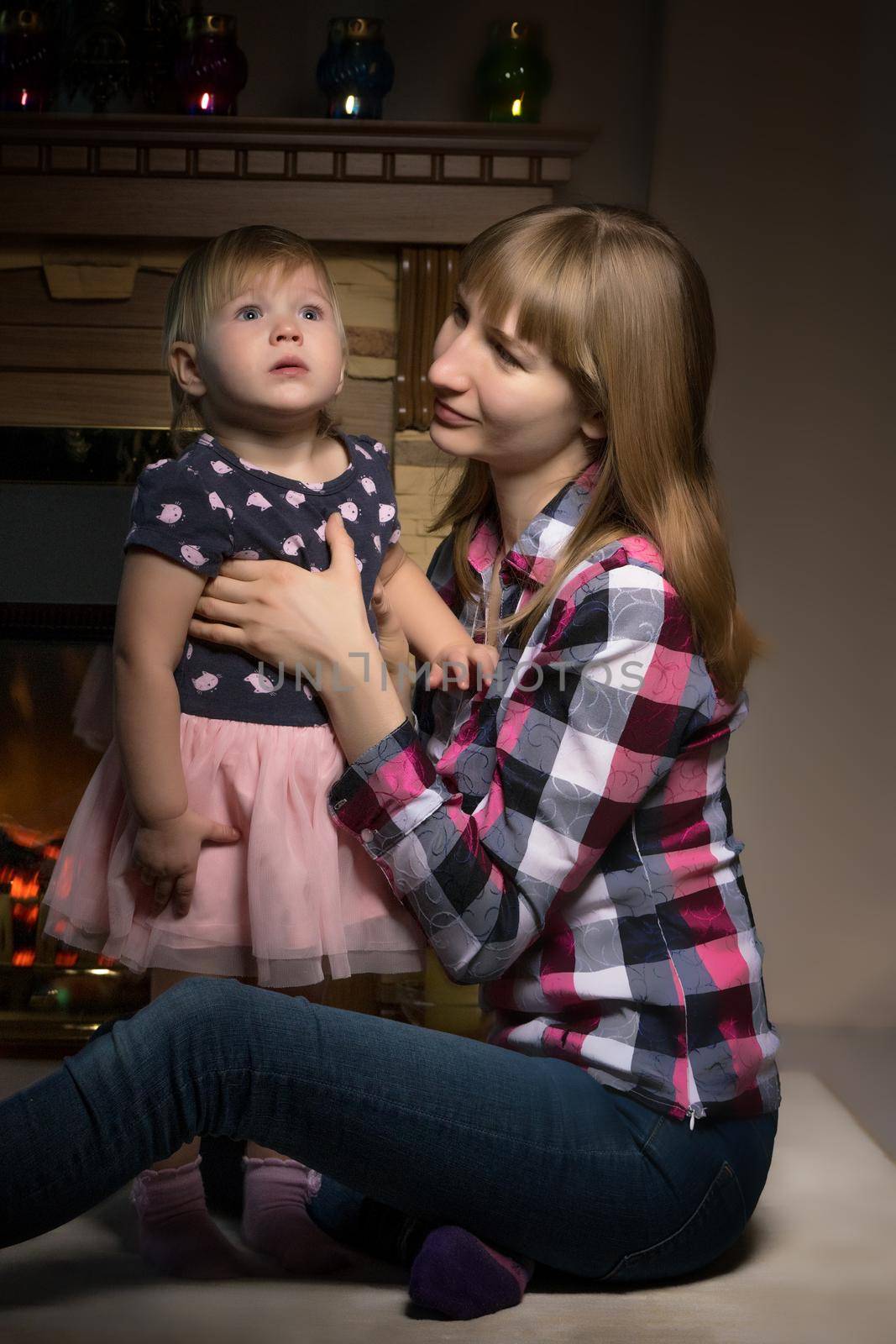 A young mother with a little girl by the fireplace on Christmas Eve. The concept of family happiness, a holiday.