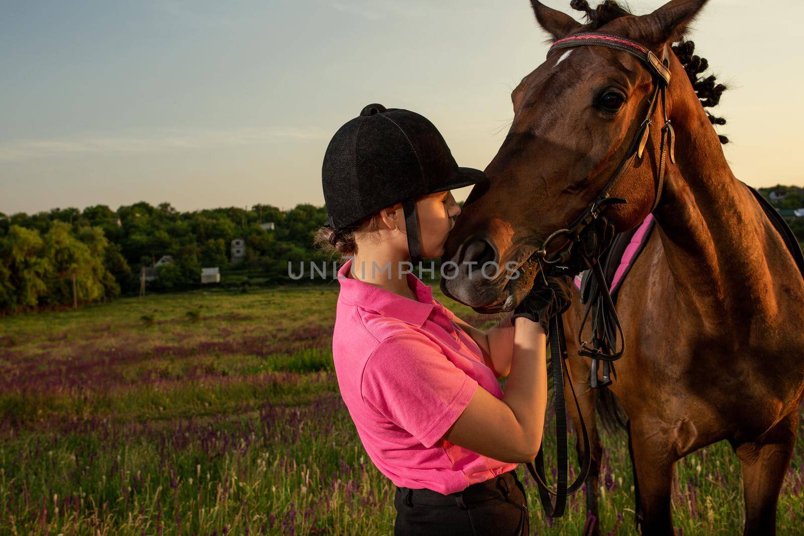 Beautiful smiling girl jockey stand next to her brown horse wearing special uniform on a sky and green field background on a sunset. by nazarovsergey