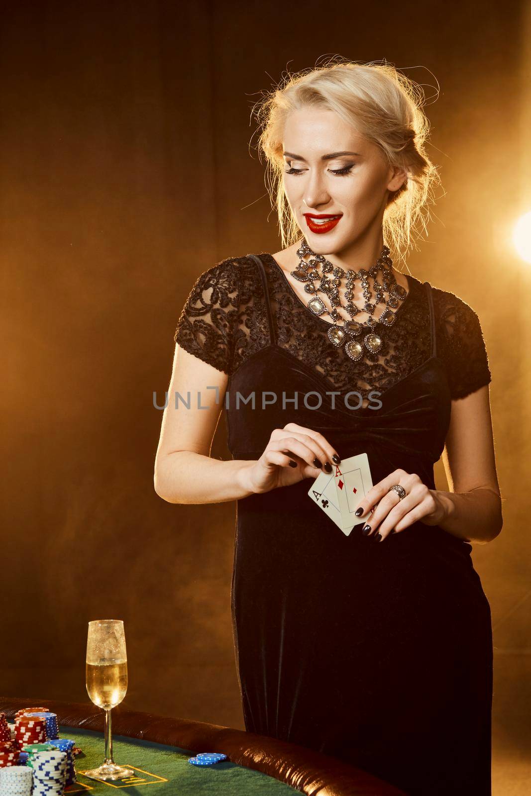 A woman in a black dress with cards in her hands is standing near the poker table. On the poker table are stacks of colored chips and a glass of champagne