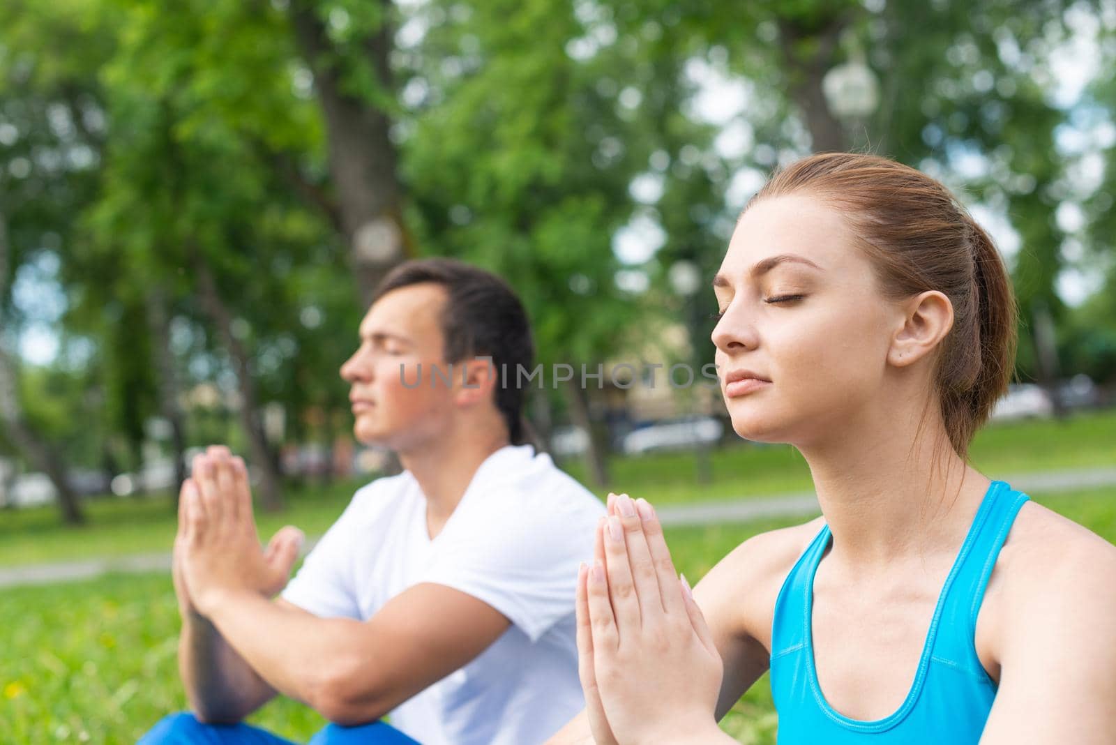 Content peaceful young couple with closed eyes sitting in lotus position. Man and woman doing yoga in park together. Meditation in yoga class. Morning exercising outdoors and healthy lifestyle.