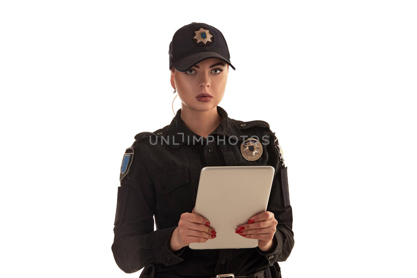 Close-up shot of a beautiful redheaded policewoman in a black uniform and a cap holding a tablet in her arms. She is looking at the camera and posing isolated on white background. Defender of citizens is ready to enforce a law and stop a crime.