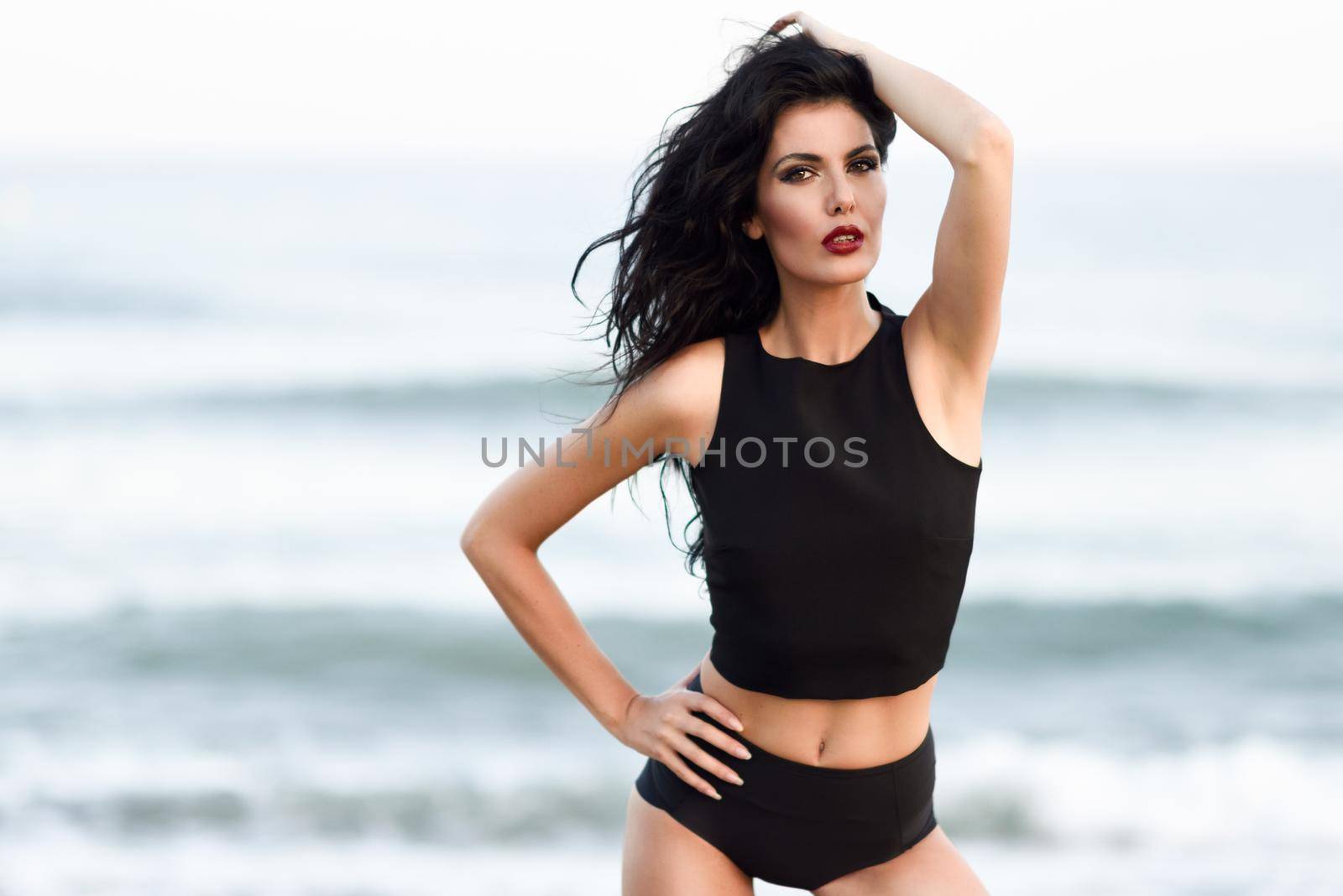 Brunette woman, model of fashion, wearing black top and panties in front of a beach. Young girl with curly hairstyle moving hair.