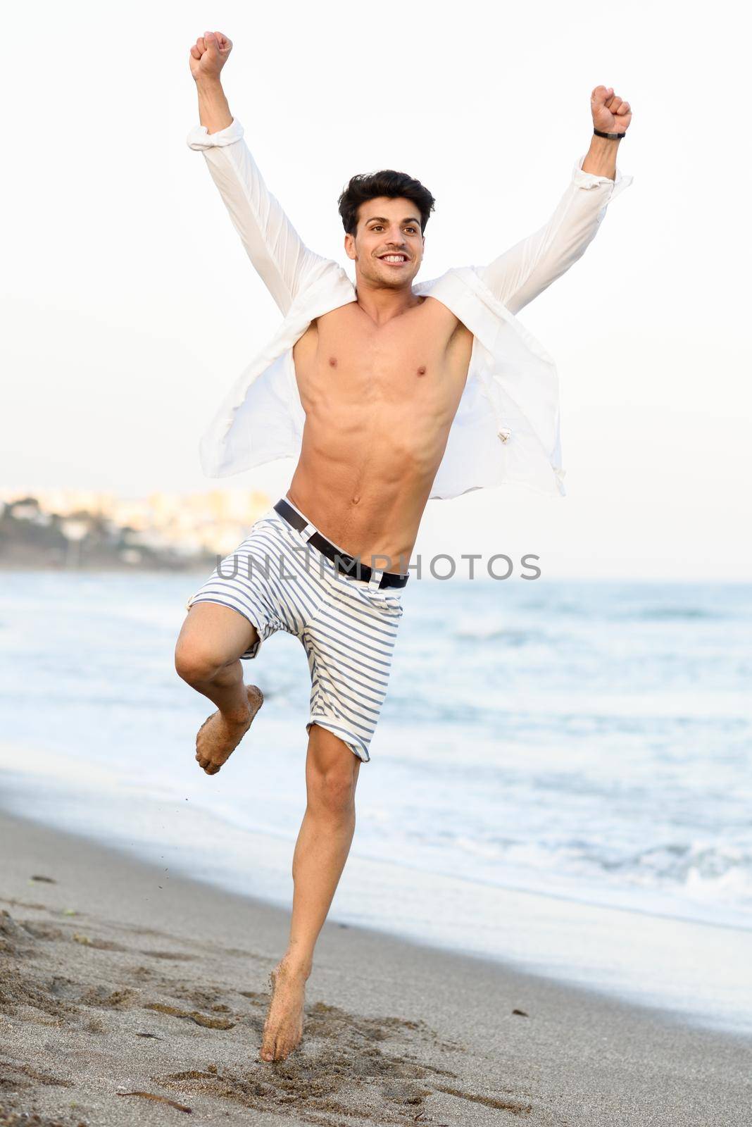 Portrait of an attractive young man jumping on a tropical beach. Handsome guy wearing white shirt and striped shorts. Male with beautiful body.