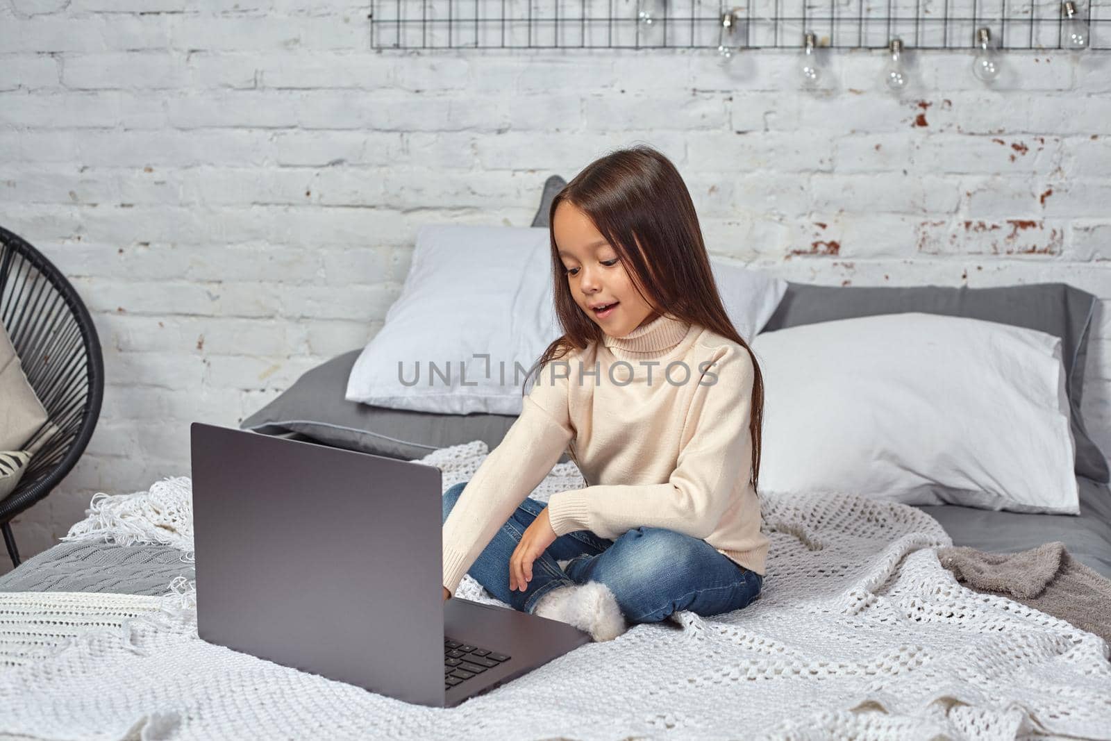 Cute little girl girl feeling amusing while watching cartoons on a laptop sitting on bed. White sweater and jeans.