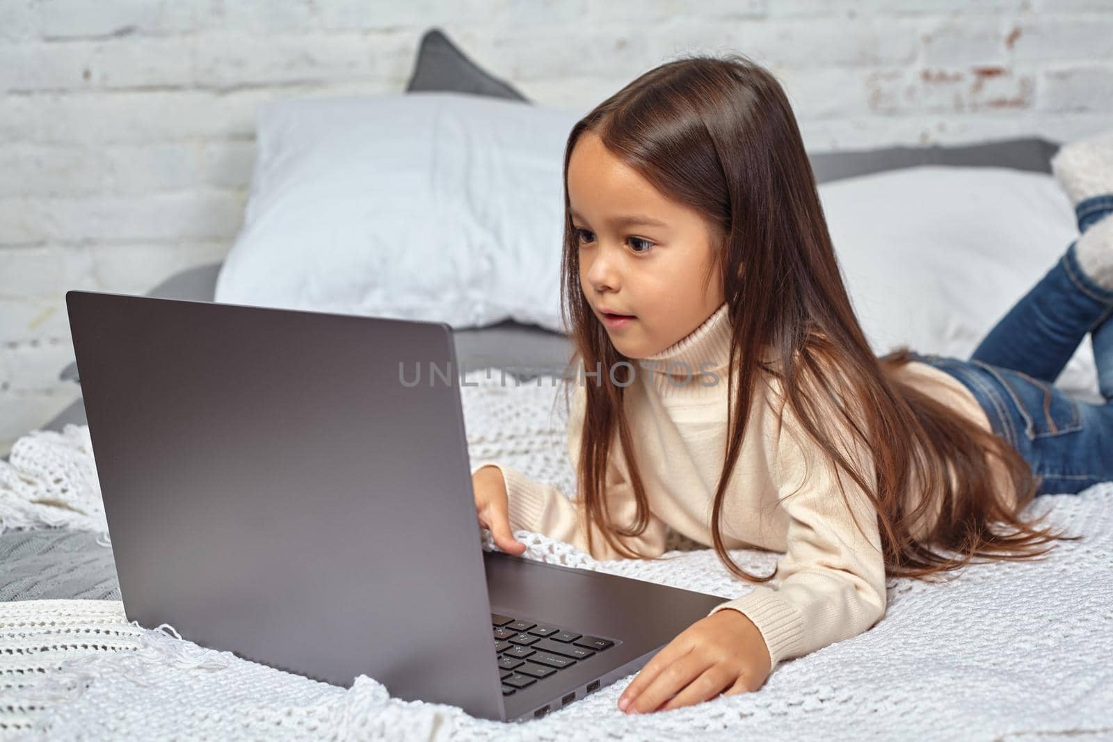 Cute little girl girl feeling amusing while watching cartoons on a laptop sitting on bed by nazarovsergey