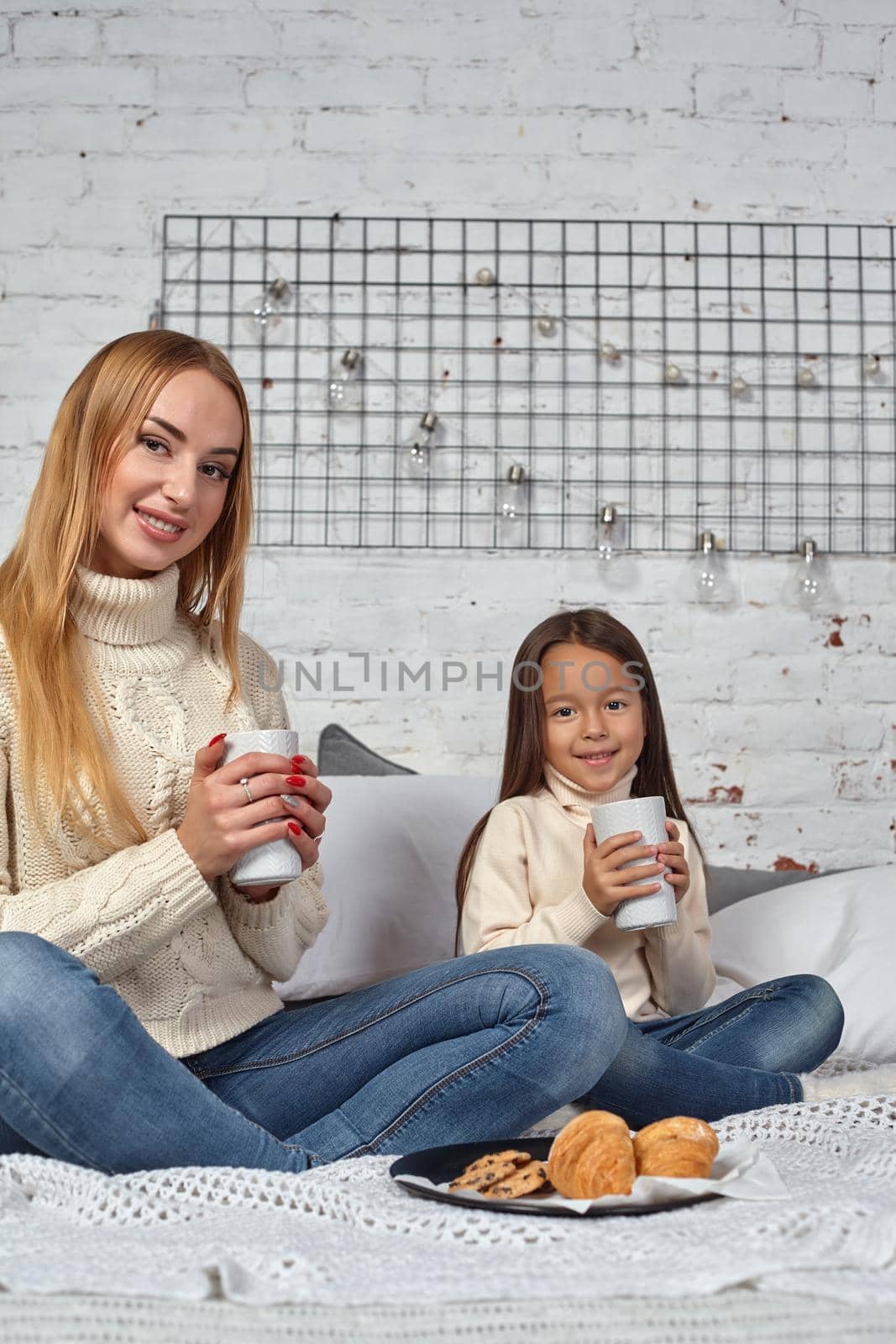 Mother and daughter in white sweaters and jeans at home, drink tea or cocoa from large mugs sitting on the bed