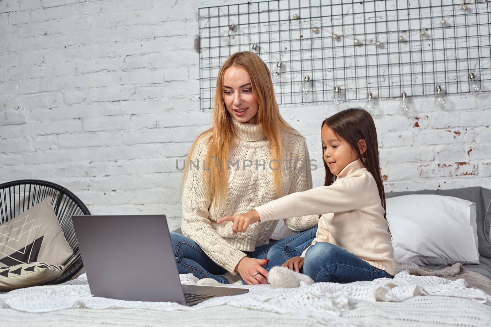 Beautiful young mother and her cute daughter in white sweaters and jeans lying on the bed at home, laughing and looking in laptop. Family time