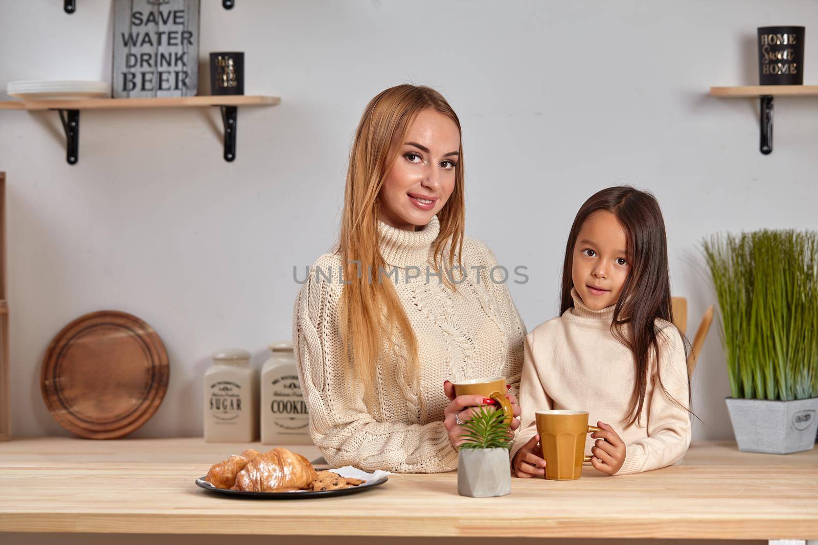 Shot of cheerful mother and daughter sit together at kitchen table, drink hot tea in morning, have pleasant friendly talk between each other. by nazarovsergey