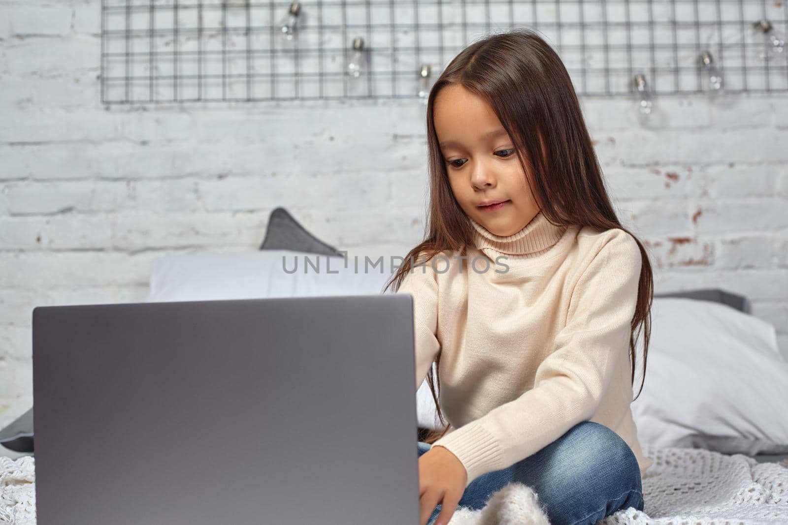 Cute little girl girl feeling amusing while watching cartoons on a laptop sitting on bed. White sweater and jeans.