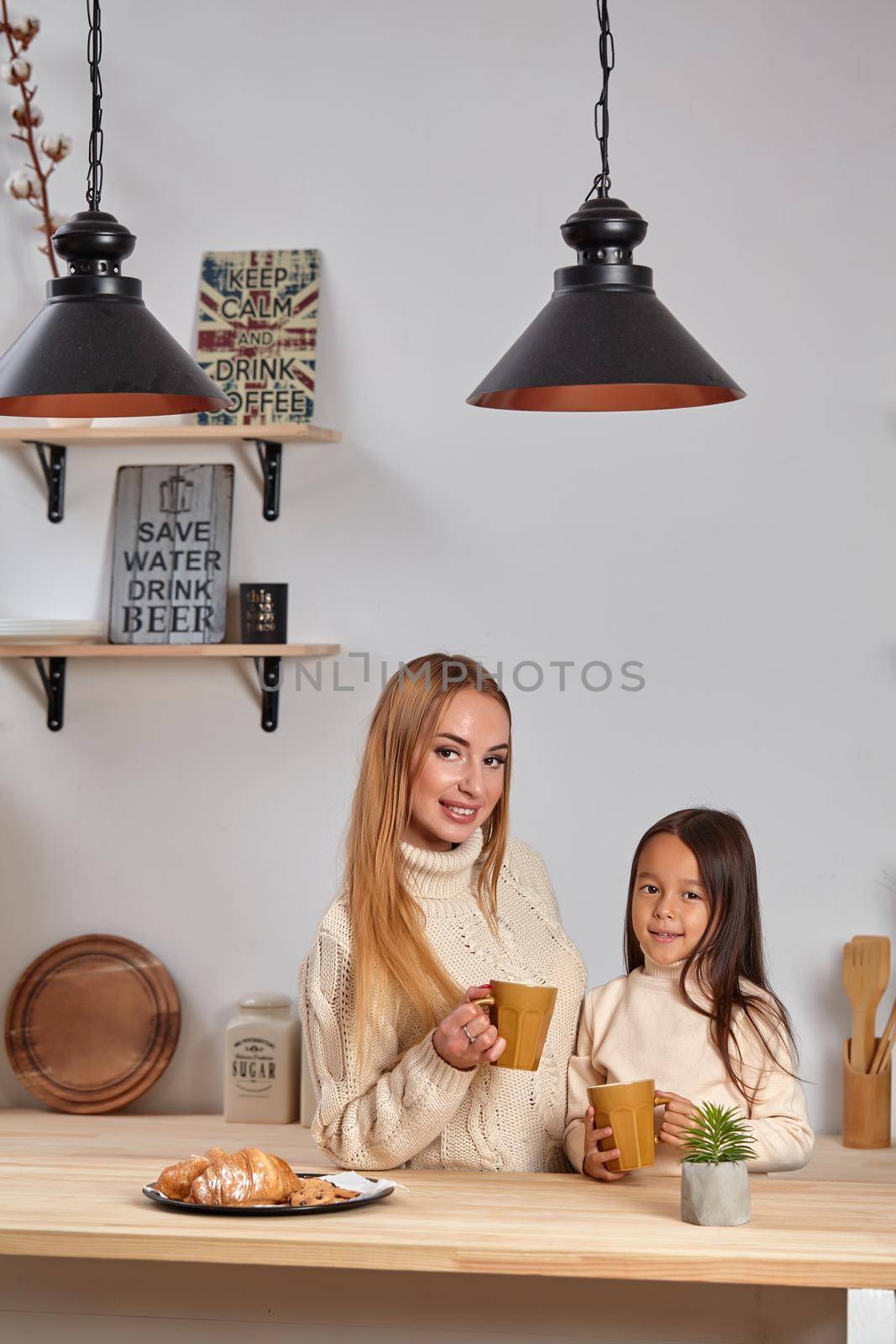 Shot of cheerful mother and daughter sit together at kitchen table, drink hot tea in morning, have pleasant friendly talk between each other. Curious girl asks something in mum during coffee break.