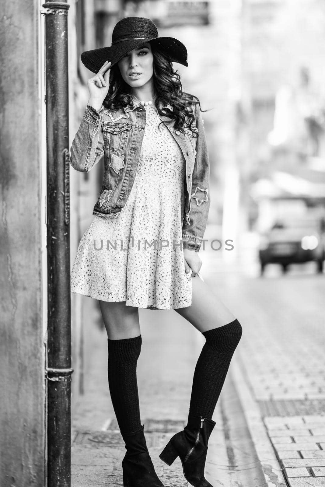Young brunette woman, model of fashion, wearing denim jacket, hat, long socks and dress. Pretty caucasian girl with long wavy hairstyle in urban background.