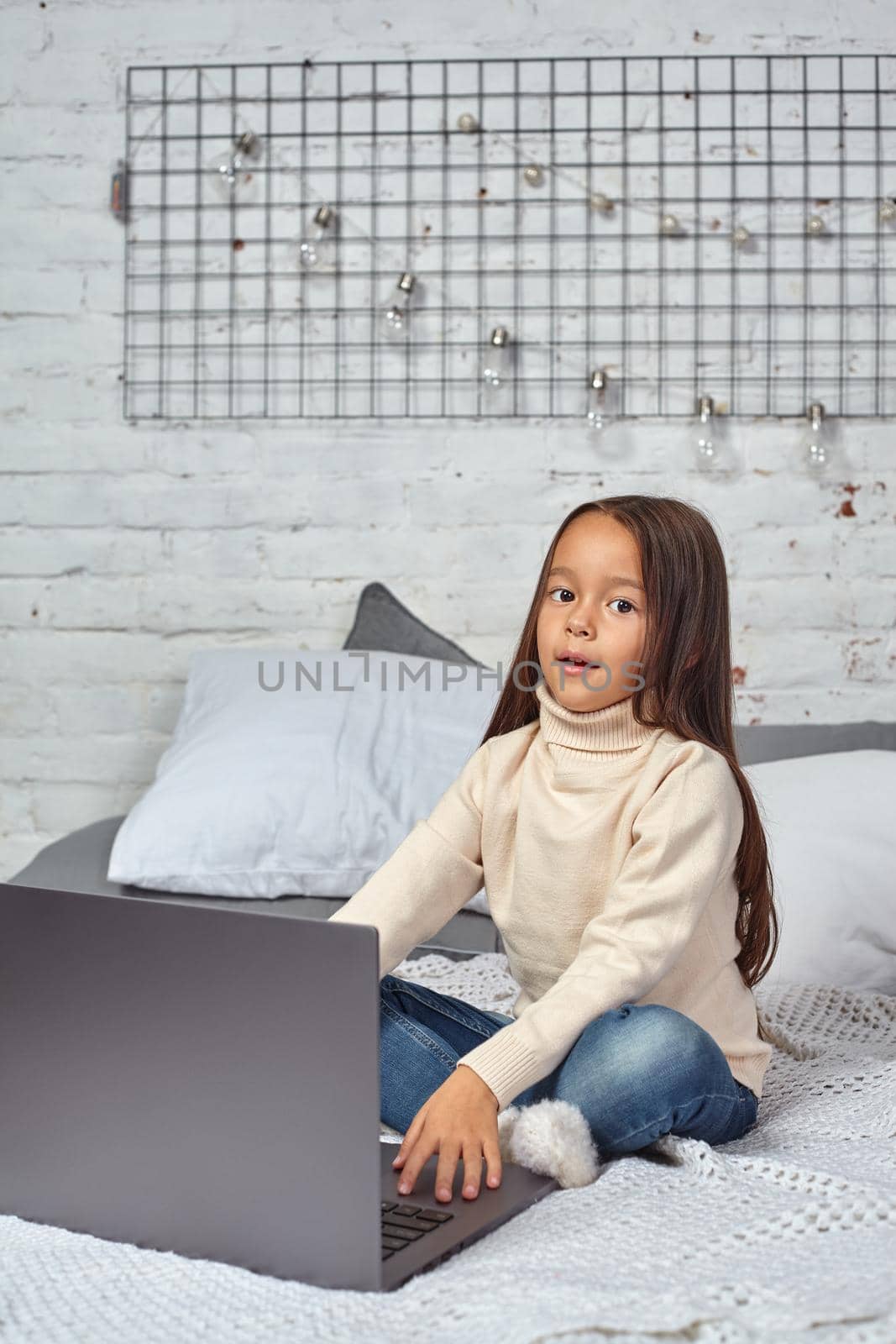 Cute little girl girl feeling amusing while watching cartoons on a laptop sitting on bed. White sweater and jeans.