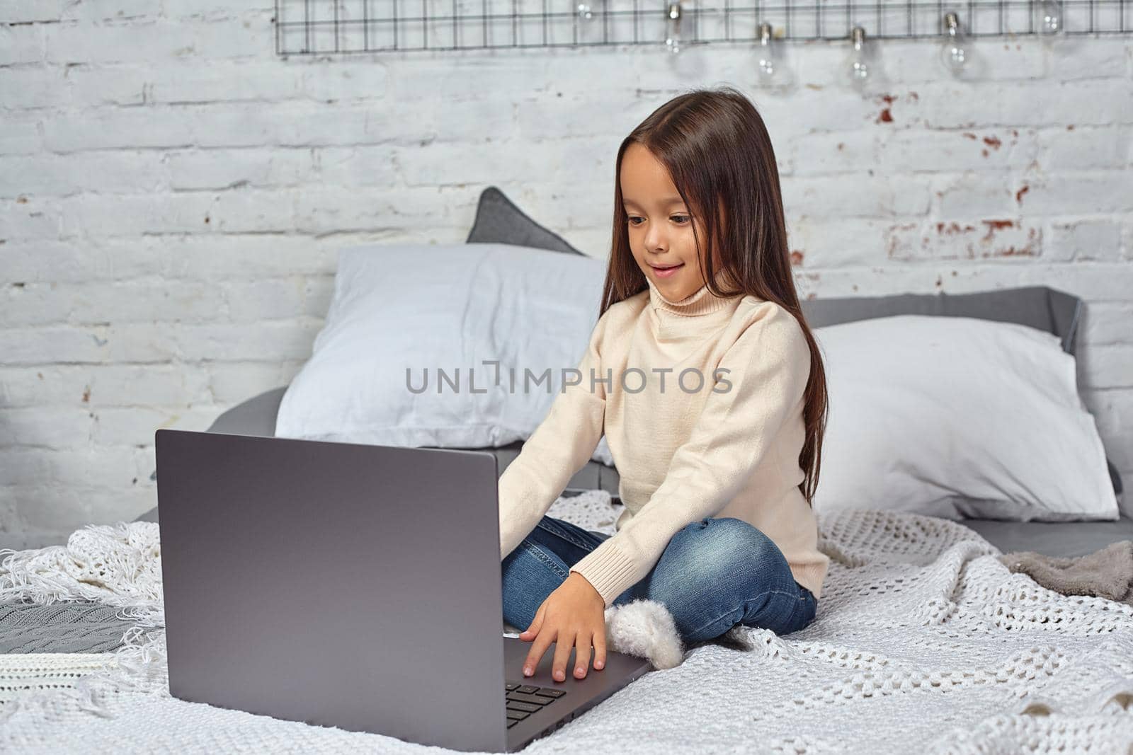 Cute little girl girl feeling amusing while watching cartoons on a laptop sitting on bed. White sweater and jeans.