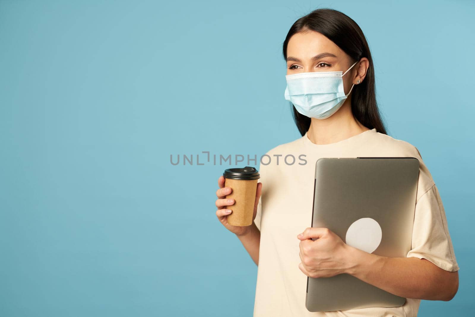 Beautiful young woman standing in studio in face mask while holding laptop and hot drink, isolated on blue background. Copy space. Quarantine, epidemic concept