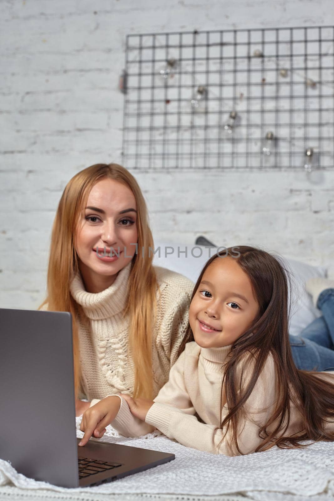 Beautiful young mother and her cute daughter in white sweaters and jeans lying on the bed at home, laughing and looking in laptop by nazarovsergey