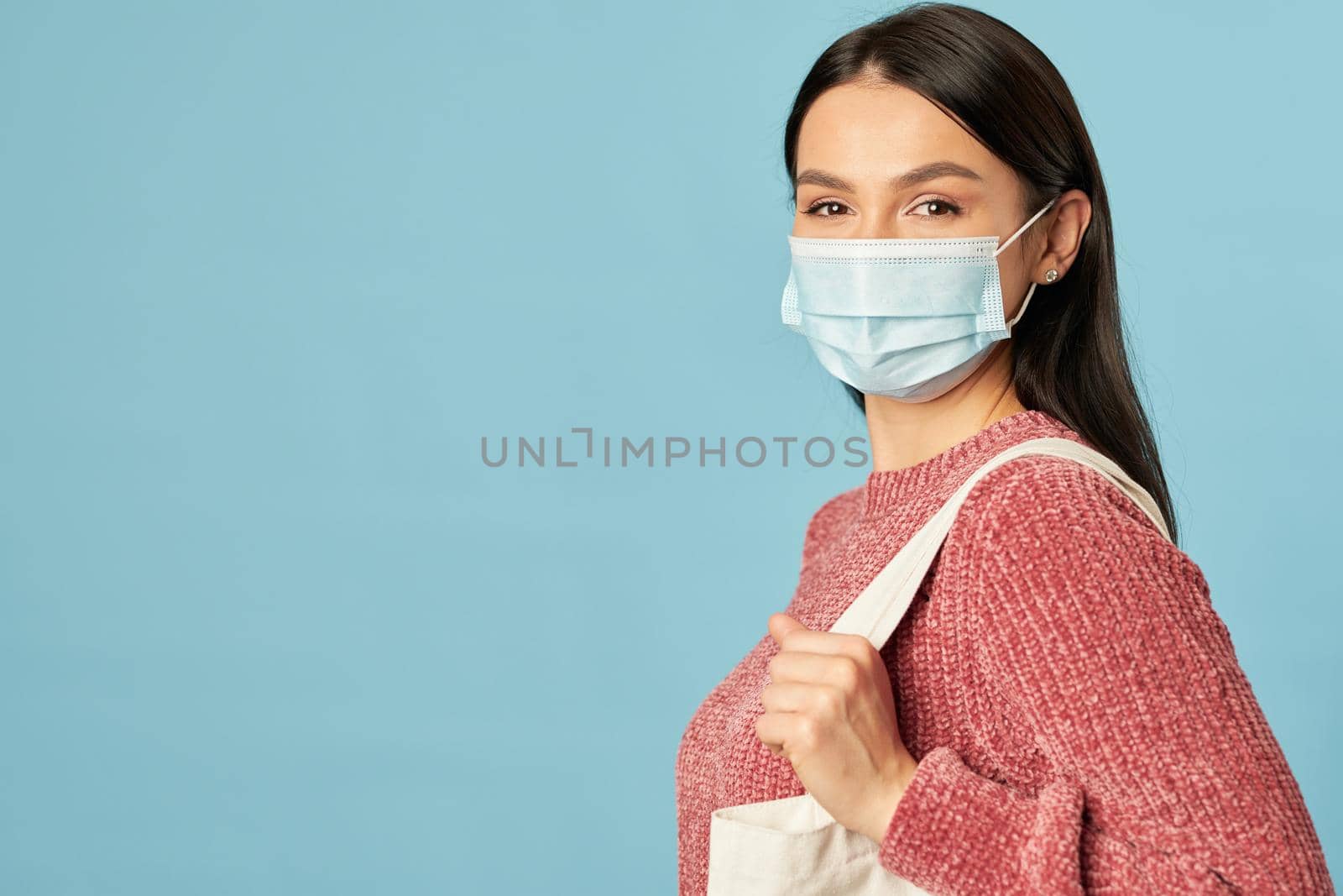 Portrait of beautiful woman in a protective face mask going shopping with a bag, isolated on blue background. Copy space. Quarantine, coronavirus concept