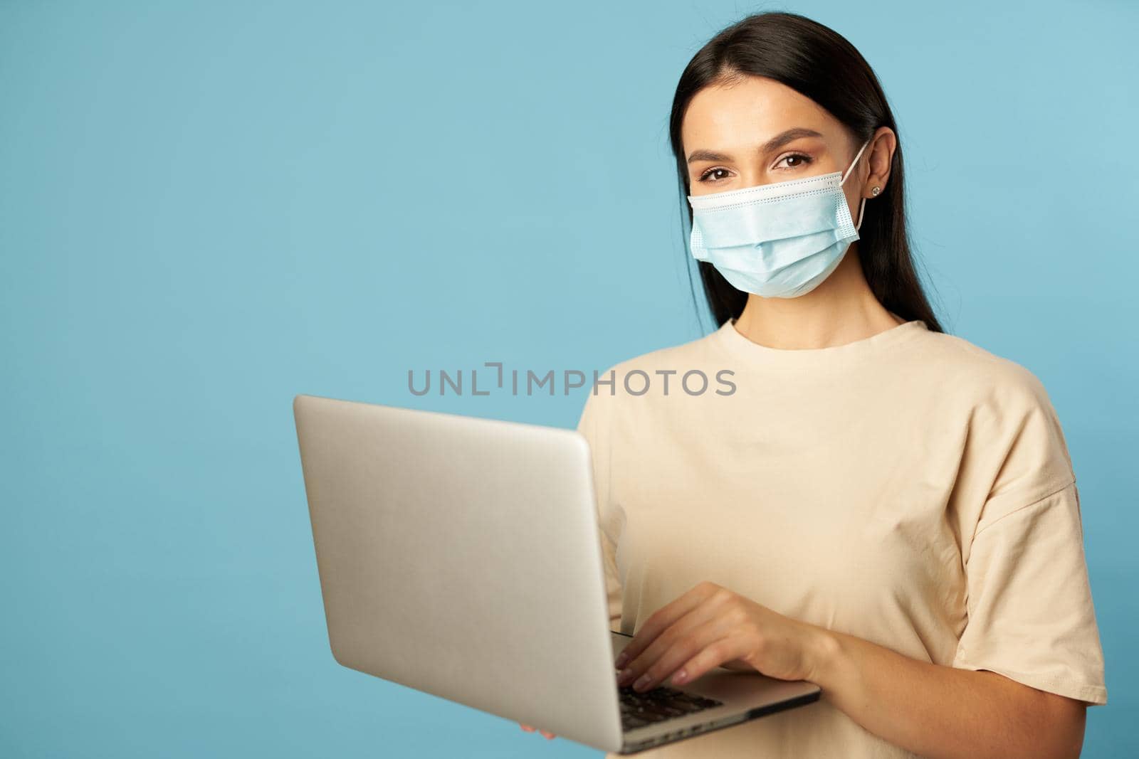 Young woman in beige t-shirt wearing protective mask and holding laptop in the studio on blue background. Copy space. Quarantine, epidemic concept