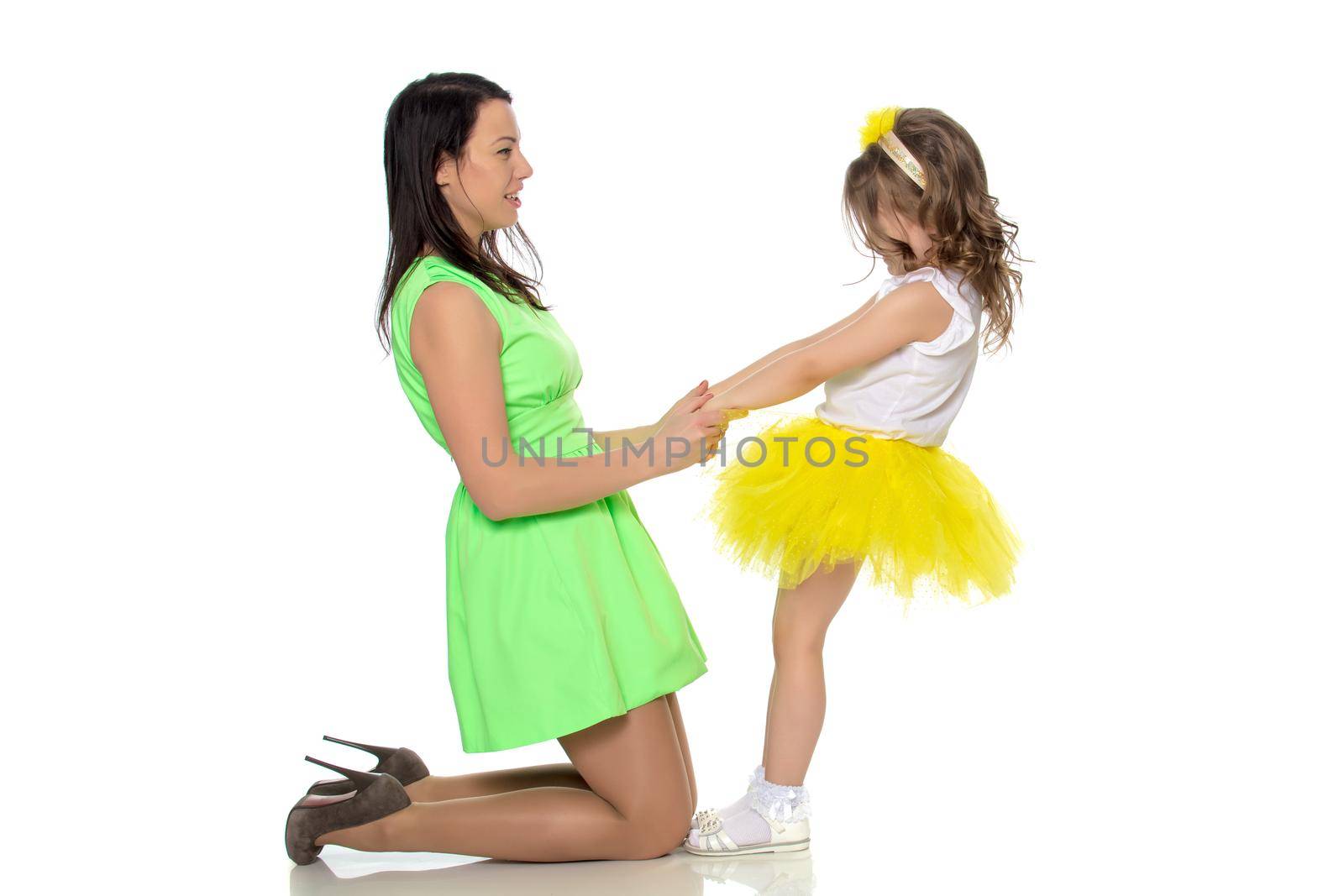 Beautiful young mother in a short green dress and her little beloved daughter in a yellow skirt.They have fun holding hands.Isolated on white background.