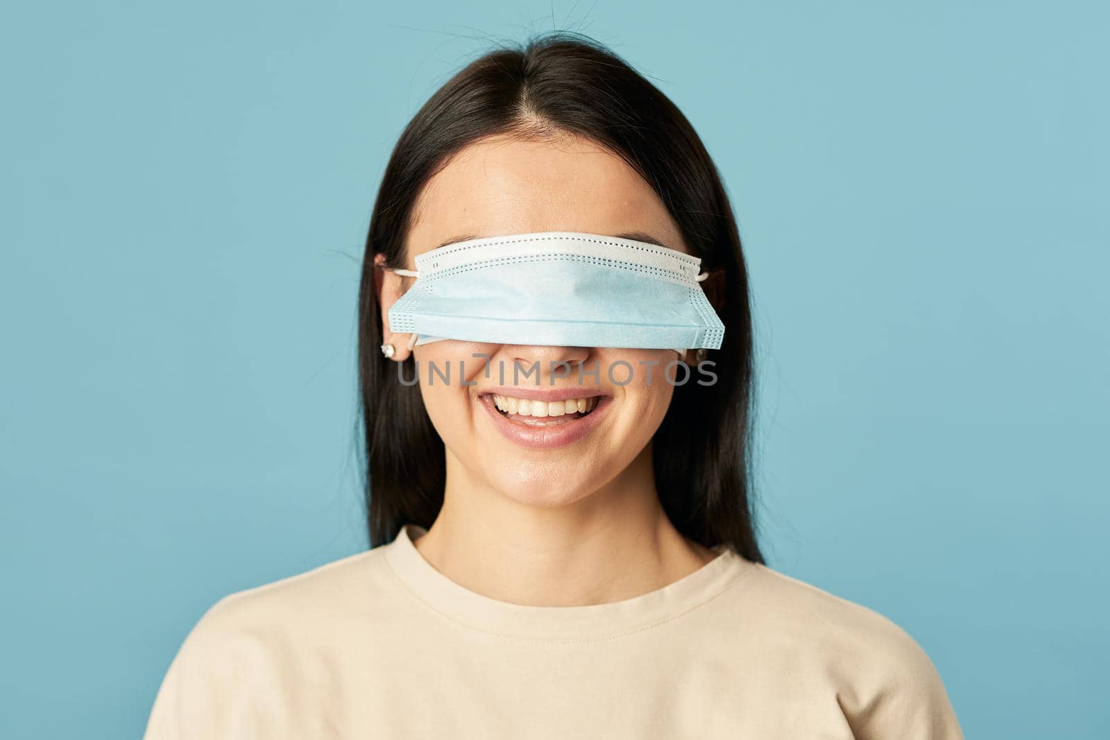 Happy woman in a beige t-shirt posing with a protective mask on her eyes on a blue background. Copy space. Quarantine, epidemic concept