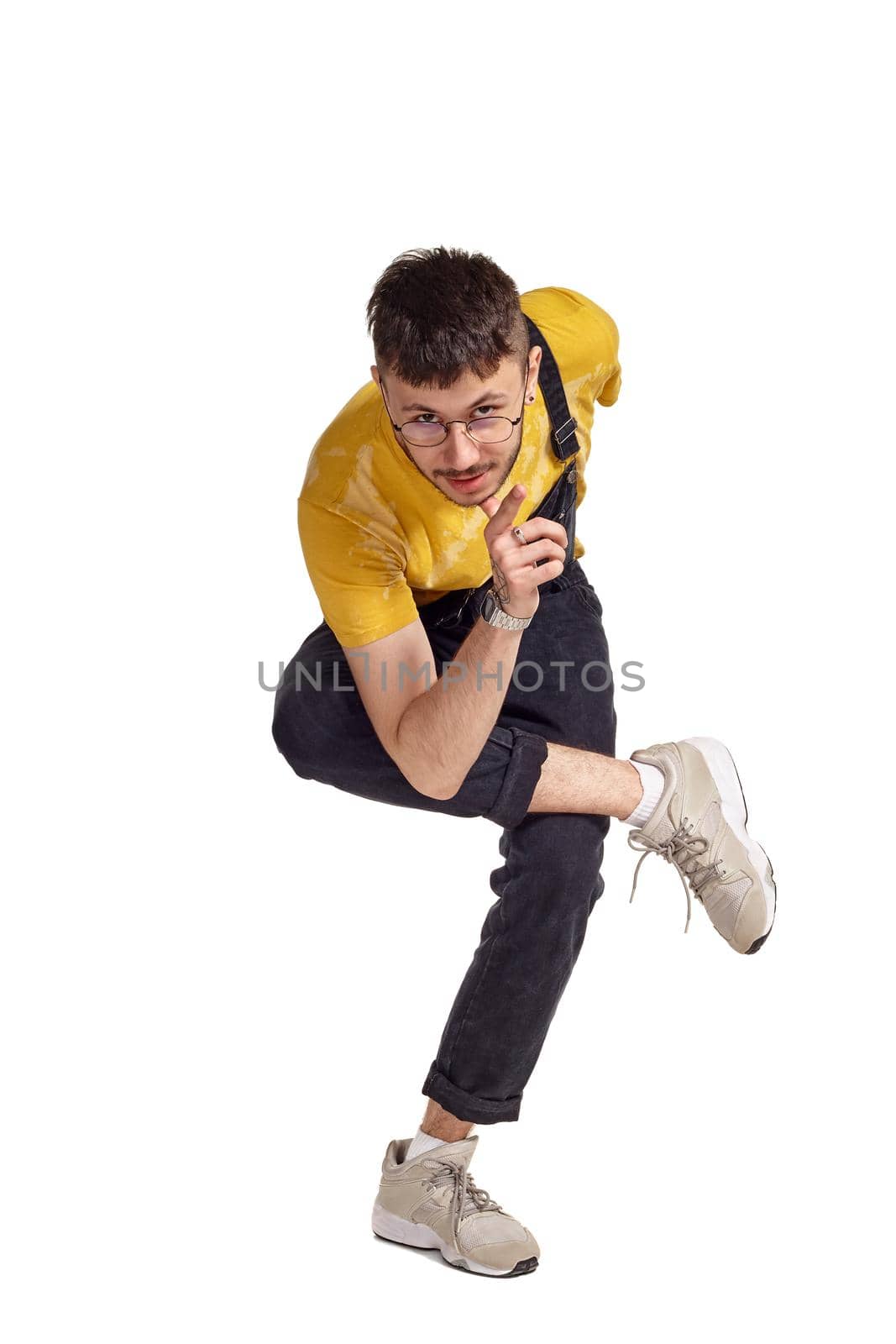 Full-length portrait of a good-looking male in glasses, black jumpsuit, yellow t-shirt and gray sneakers fooling around in studio. Indoor photo of a man making dancing elements isolated on white background. Music and imagination.