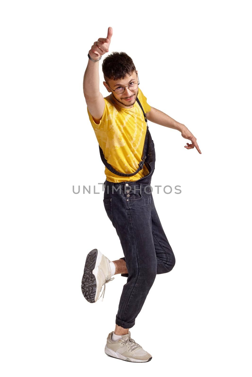 Full-length portrait of an athletic guy in glasses, black jumpsuit, yellow t-shirt and gray sneakers fooling around in studio. Indoor photo of a man making dancing elements raising his index finger isolated on white background. Music and imagination.