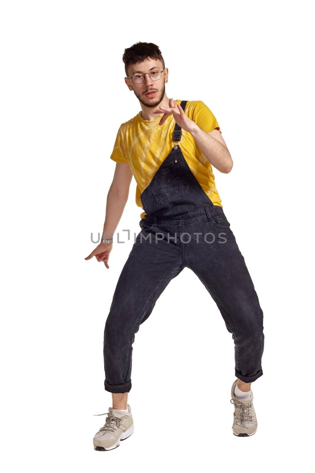 Full-length portrait of a creative person in glasses, black jumpsuit, yellow t-shirt and gray sneakers fooling around in studio. Indoor photo of a man making dancing elements isolated on white background and looking at the camera. Music and imagination.