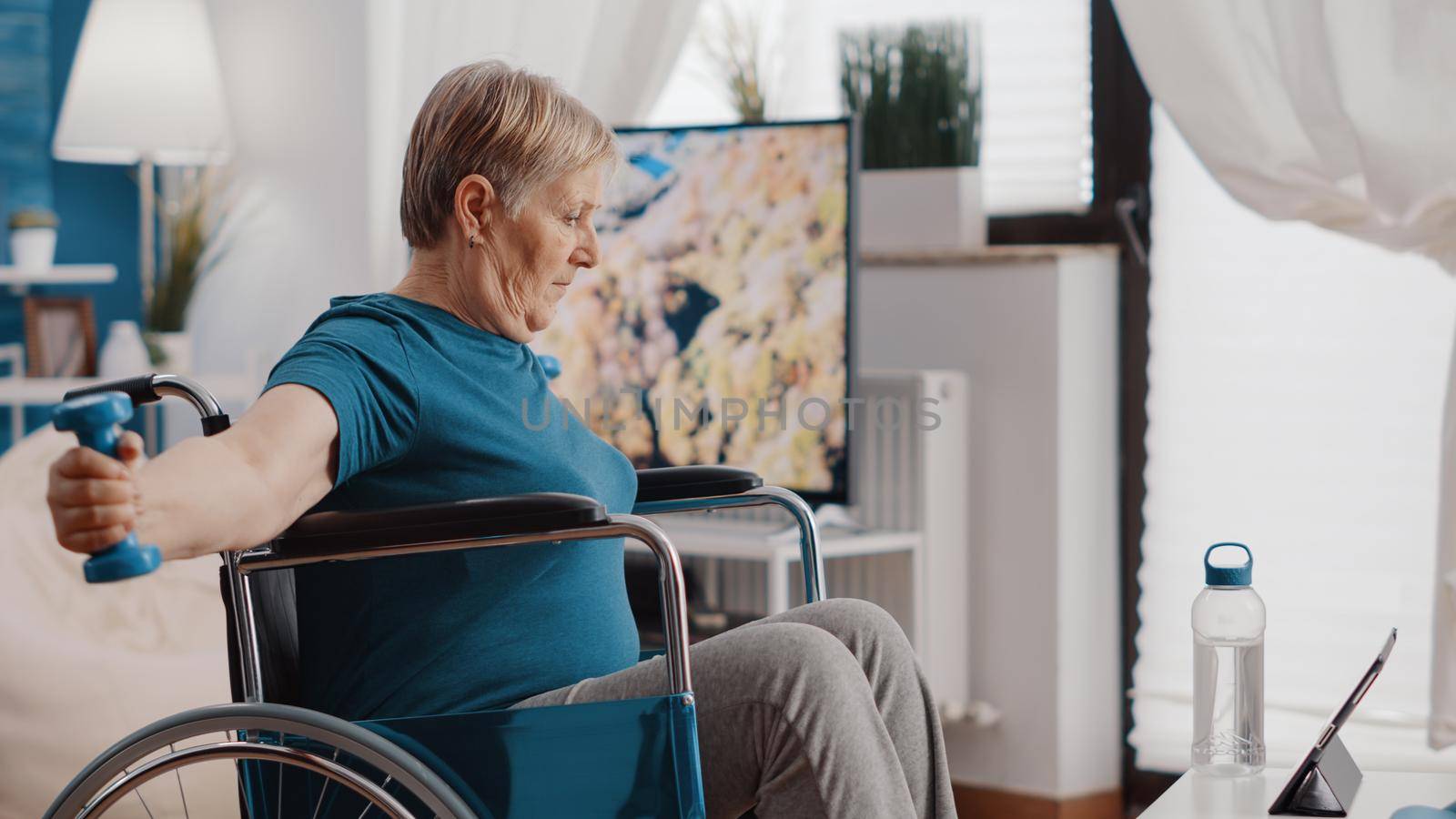 Old woman with disability using dumbbells to exercise and watching workout video on digital tablet. Retired person sitting in wheelchair, training with weights and looking at online lesson