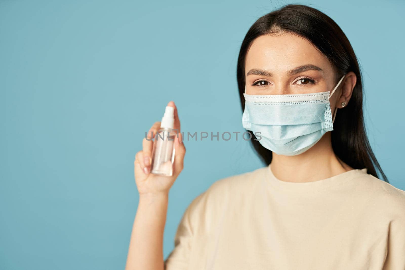 Pretty lady in a protective face mask posing in the studio and holding an antiseptic on blue background. Quarantine, coronavirus concept