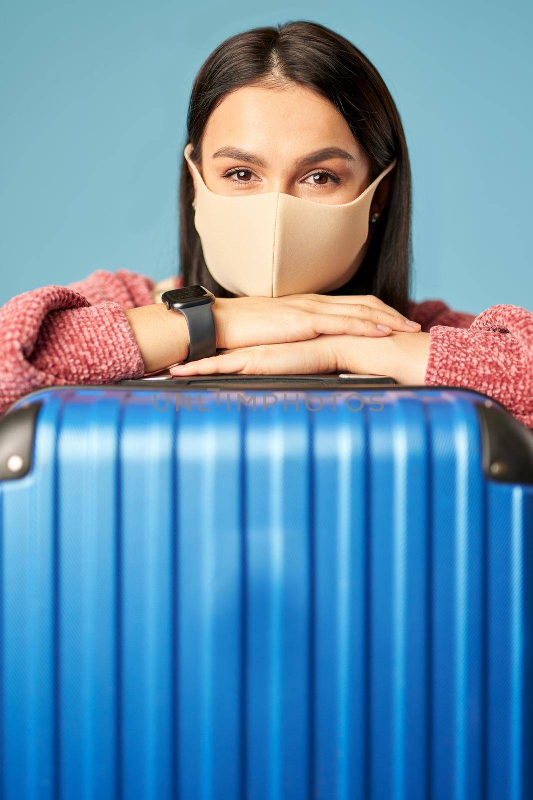Beautiful lady in a sweater with a suitcase posing in studio, isolated on blue background. Copy space. Concept of travel, coronavirus