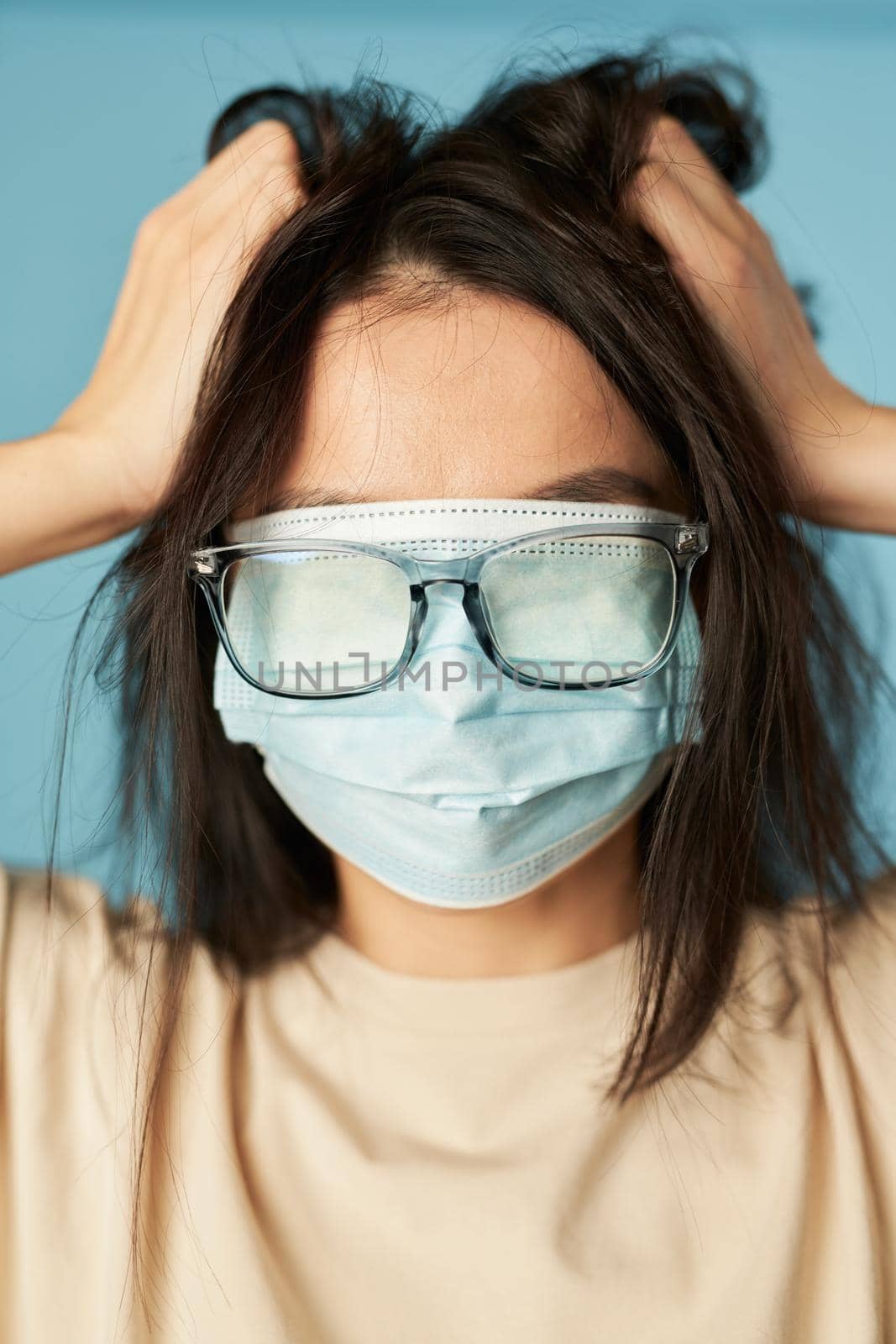 Close up of lady with tousled hair posing with a protective mask and glasses on her eyes on a blue background. Copy space. Quarantine, epidemic concept