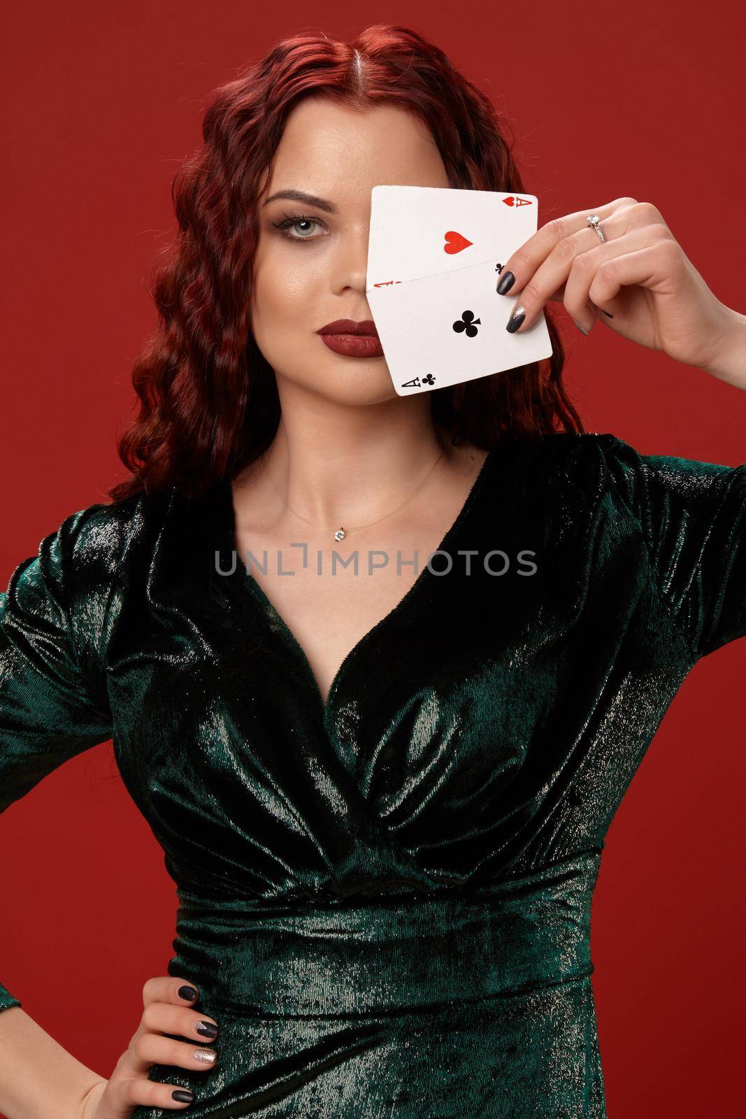 Young sexy woman with a red curly hair, have brought playing cards to her face and looking at the camera, on a red background. Poker