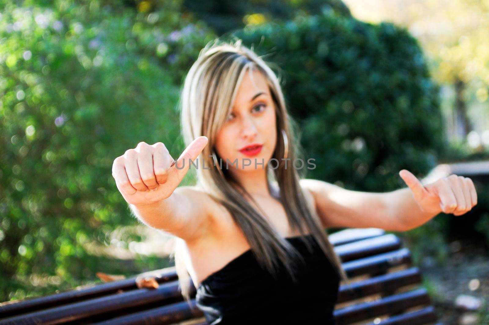 Pretty girl sitting in a bench in the park showing thumb up sign by javiindy