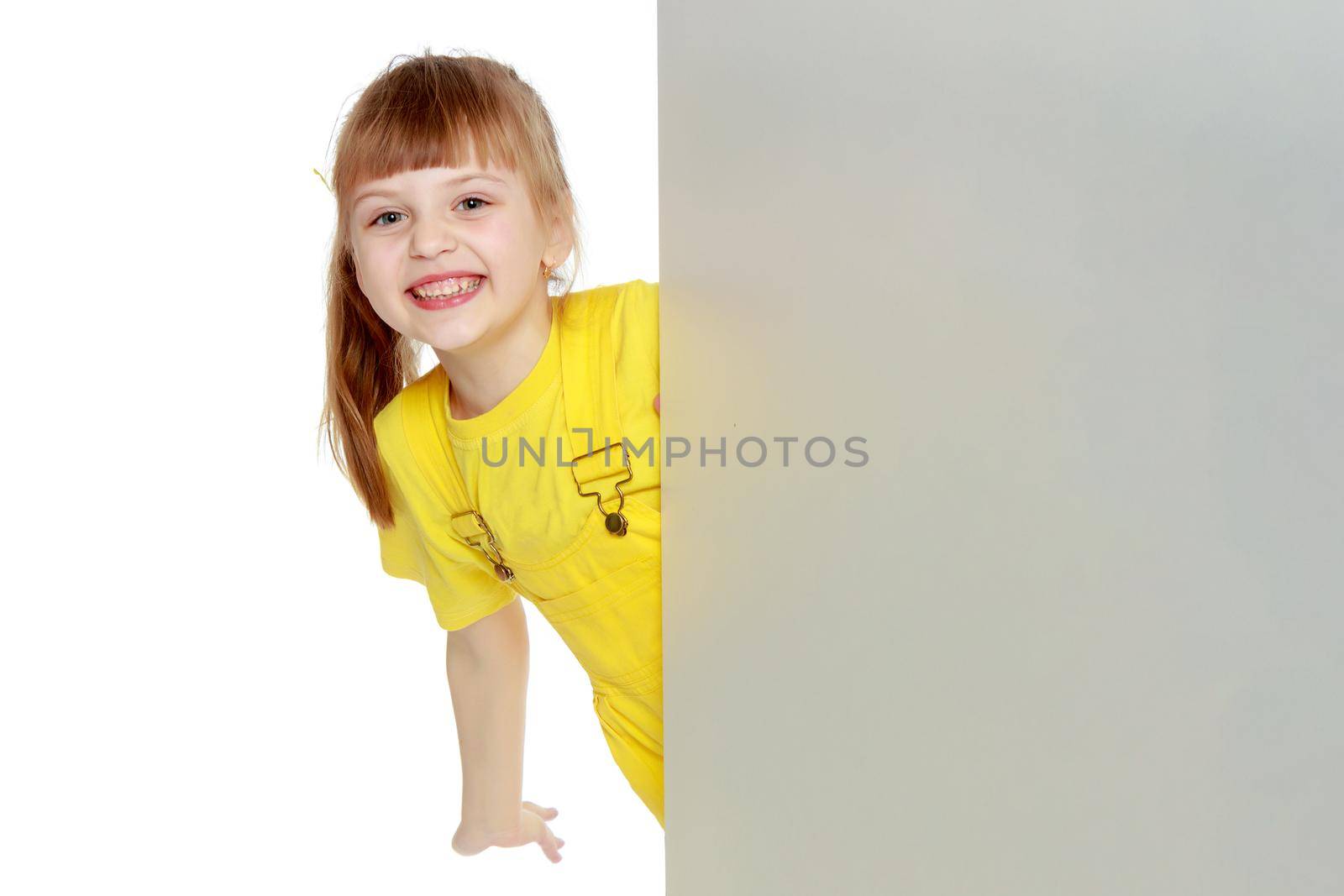 Girl with a short bangs on her head and bright yellow overalls.She crouched down on the white advertising banner.