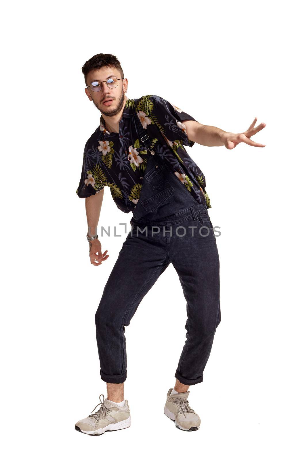 Full-length portrait of a handsome dancer in glasses, black jumpsuit, colorful t-shirt and gray sneakers fooling around in studio. Indoor photo of a man dancing isolated on white background and looking at the camera. Music and imagination.