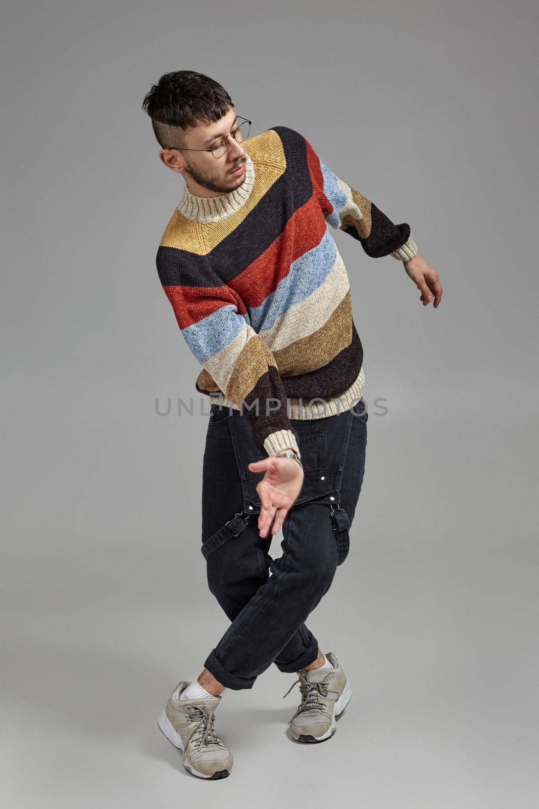 Full-length photo of a handsome male in glasses, black jumpsuit, multi-colored sweater and gray sneakers fooling around in studio. Indoor portrait of a man dancing on a gray background. Music and imagination.