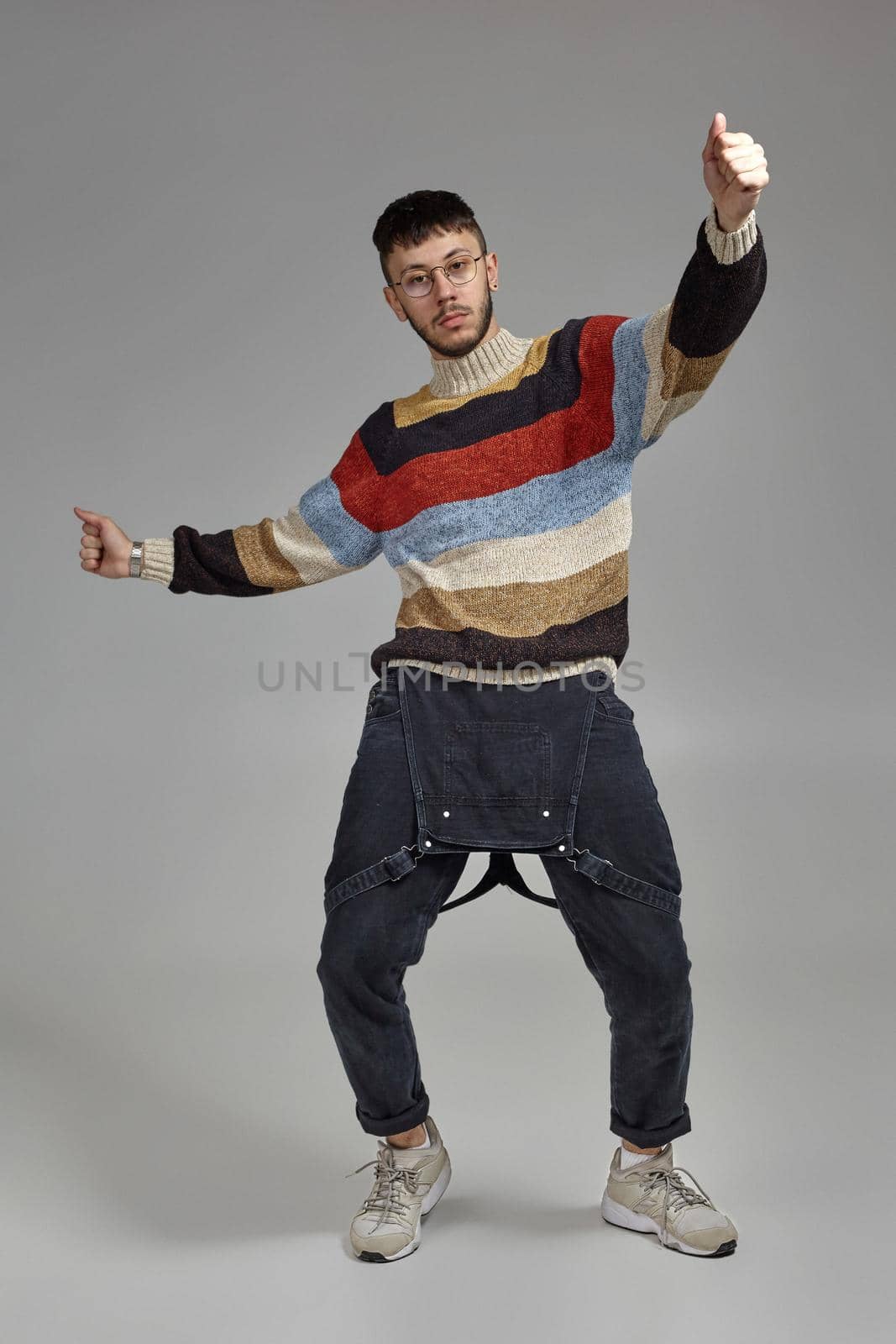 Full-length portrait of a funny man in glasses, black jumpsuit, multi-colored sweater and gray sneakers fooling around in studio. Indoor photo of a guy dancing and looking at the camera on a gray background. Music and imagination.