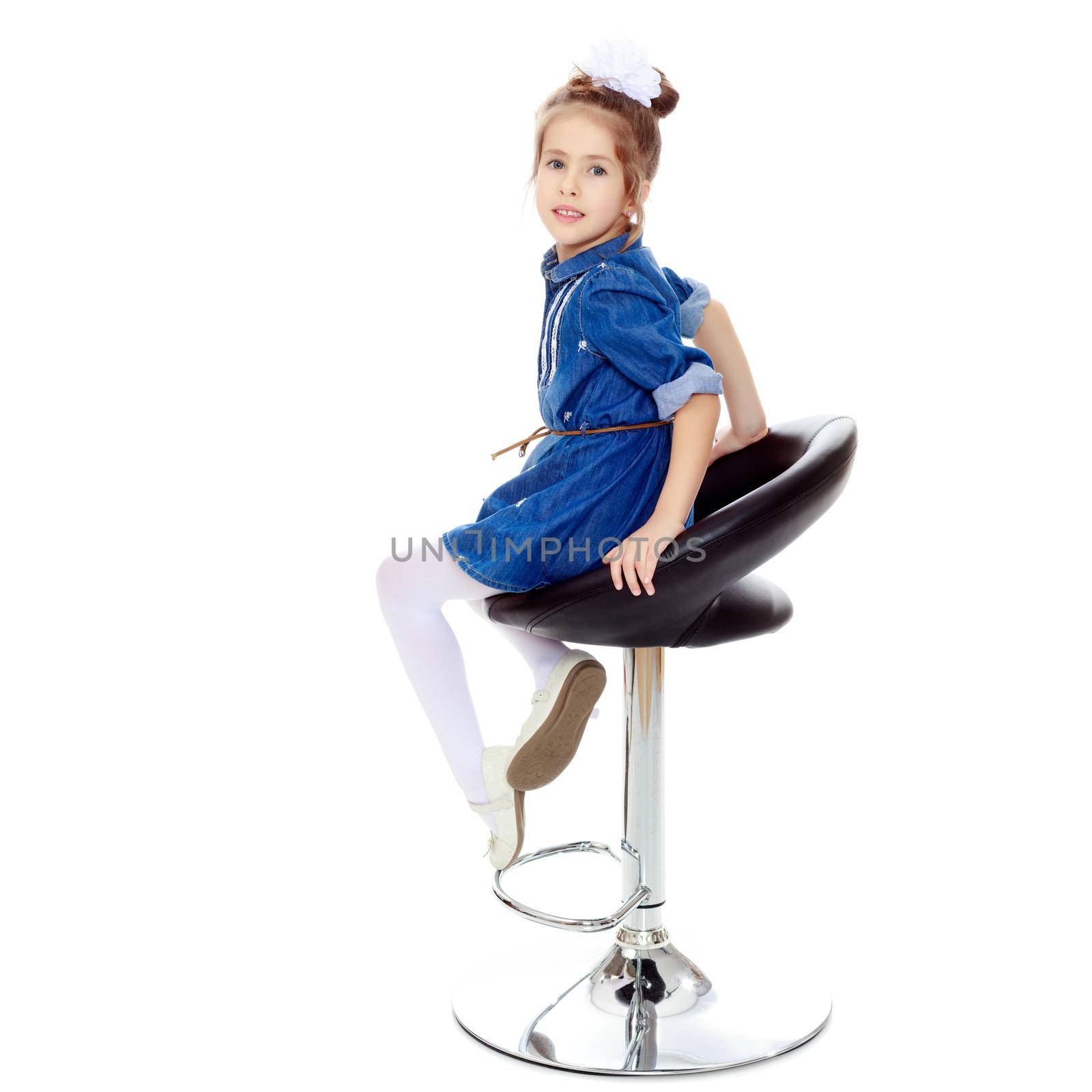 The little blonde girl with a large white bow on the head and short denim dress.She poses on a revolving chair.Isolated on white background.
