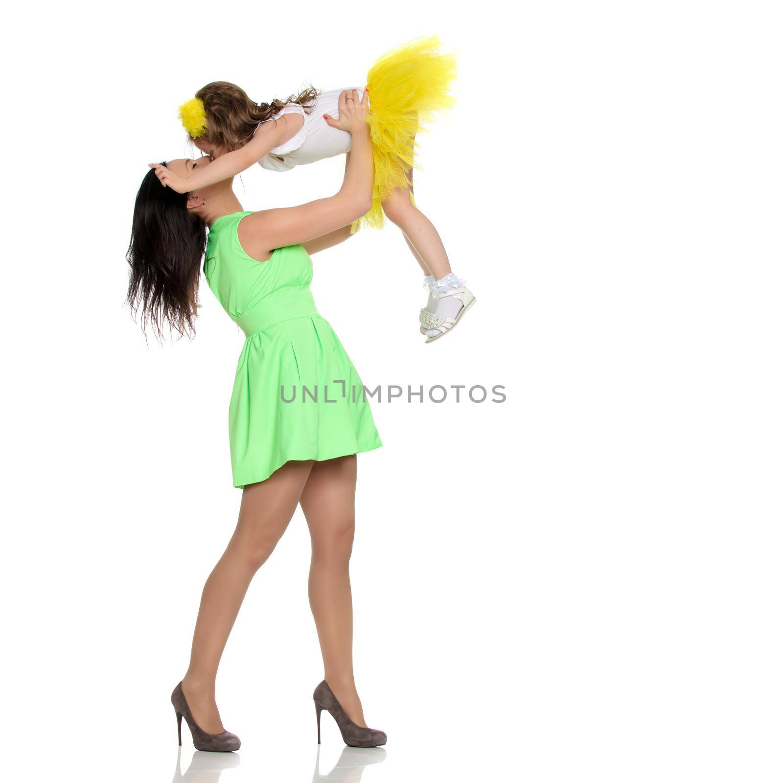 Beautiful young mother in a short green dress and her little beloved daughter in a yellow skirt.Fun mother throws daughter on hands.Turning sideways to the camera.Isolated on white background.