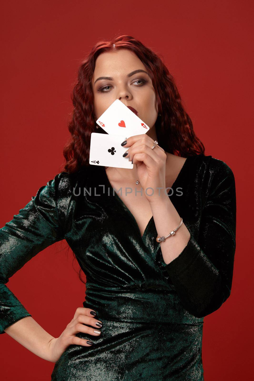 Young sexy woman with a red curly hair, have brought playing cards to her face and looking away, on a red background. Poker