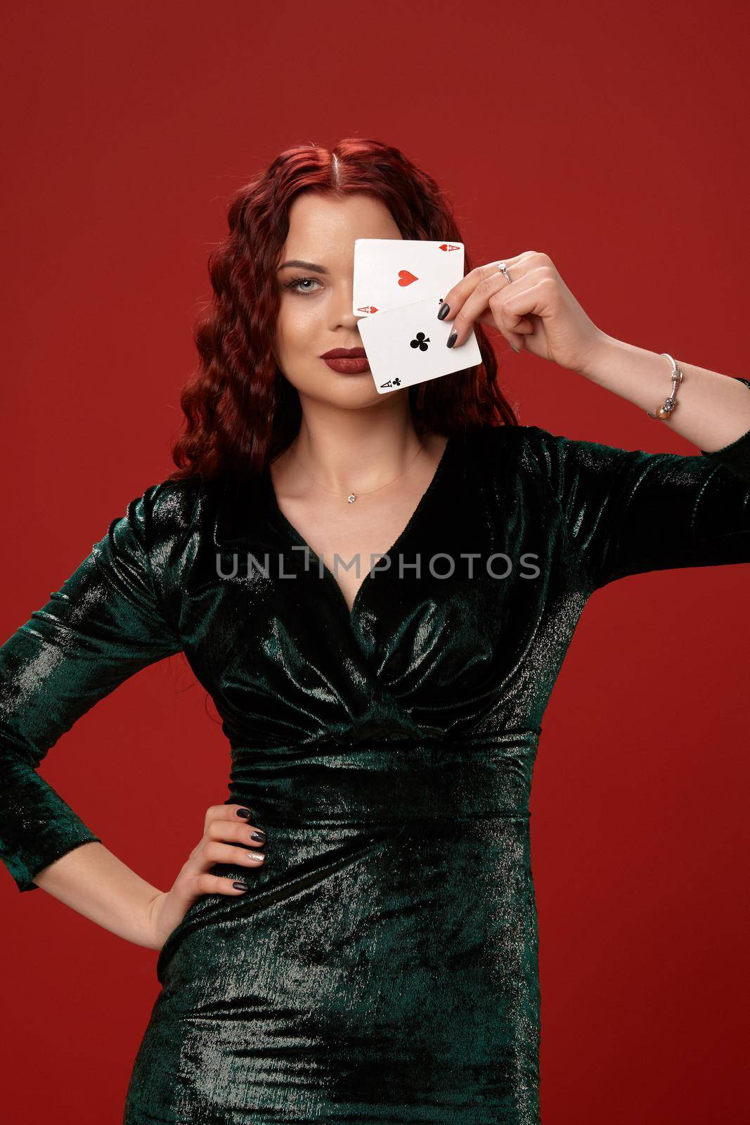 Young sexy woman with a red curly hair, have brought playing cards to her face and smiling at the camera, on a red background. Poker